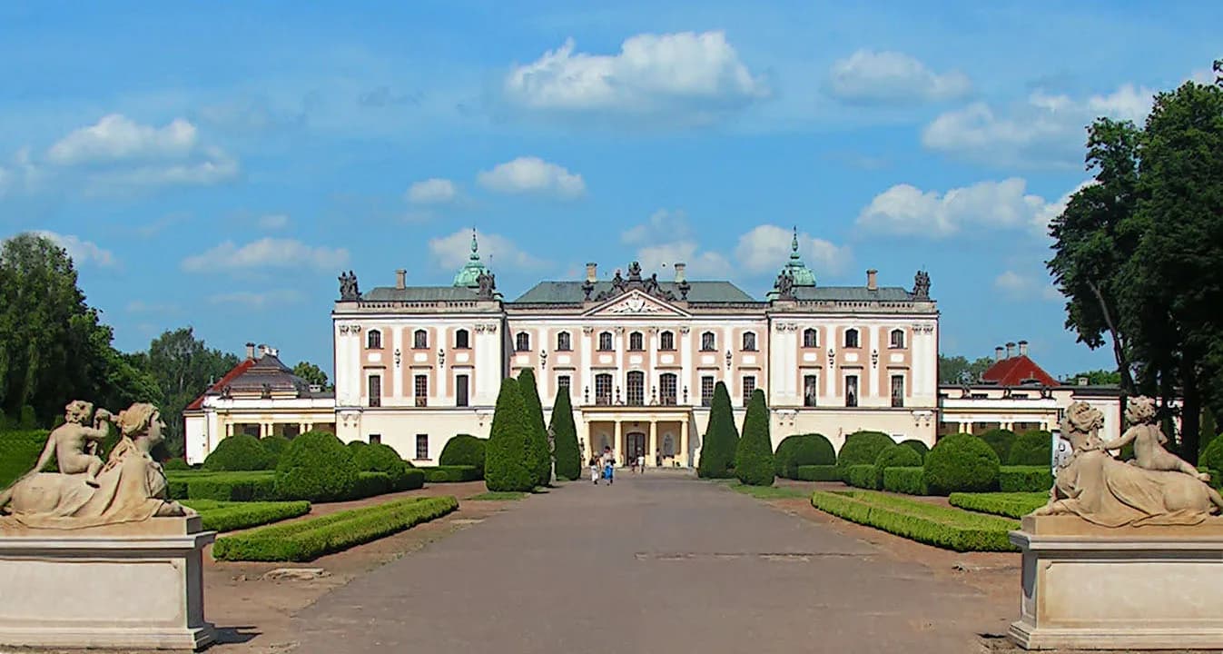 A grand, symmetrical white and beige palace with green domed roofs, surrounded by manicured green hedges and trees under a partly cloudy blue sky, with a paved pathway leading up to its entrance and stone statues on pedestals in the foreground.